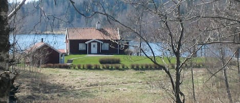 Una casa con techo rojo, puerta blanca y un pequeño porche. Se observa una masa de agua al frente y un área boscosa al fondo.