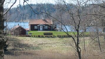 A house with a red roof, a white door, and a small porch. There is a body of water in the foreground and a forested area in the background.