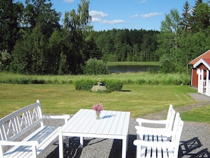 A white outdoor seating area with a table and benches, surrounded by a grassy field and a pond, with a red building in the background. - Holiday Home in Åtvidaberg (Atvidaberg)