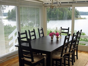 A dining table with six chairs, a chandelier, and a view of a lake through large windows with blinds. - Holiday Home in Åtvidaberg (Atvidaberg)