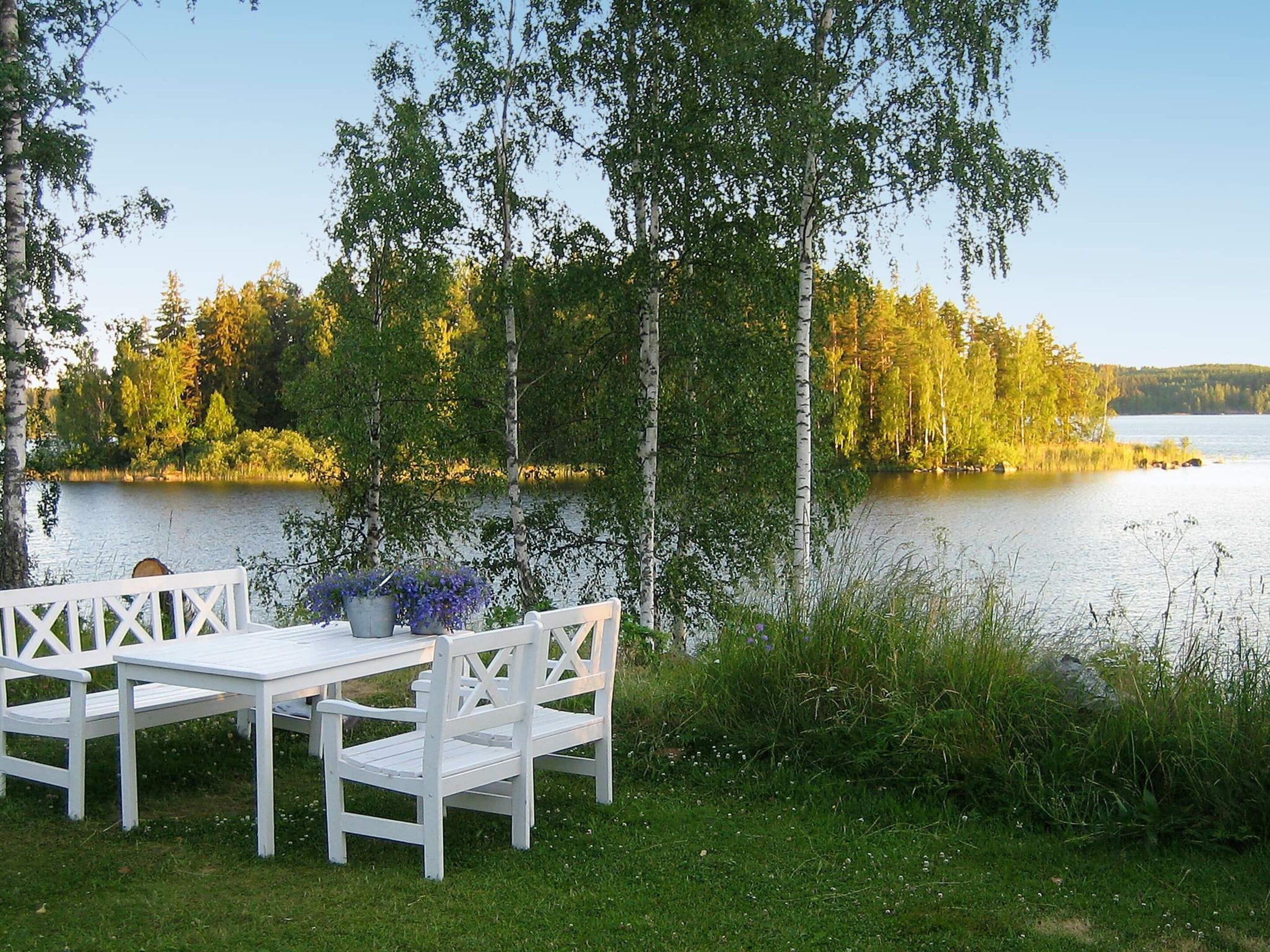 A lakeside setting with a white wooden bench and table, surrounded by trees and a calm body of water.
