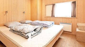 Una habitación con paredes de madera, una cama con almohadas y mantas, una ventana con cortinas, un radiador y una cesta de mimbre.