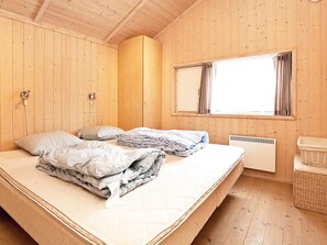A room with wooden walls, a bed with pillows and blankets, a window with curtains, a radiator, and a wicker basket. - 4 Star Holiday Home in Rodby (Rodby)