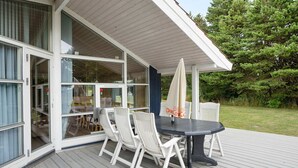 A covered outdoor deck with a table and chairs, large windows, and a view of the surrounding greenery. - 4 Star Holiday Home in Rodby (Rodby)