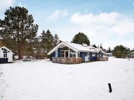Una casa de un solo piso con techo a dos aguas, rodeada de nieve y árboles altos.