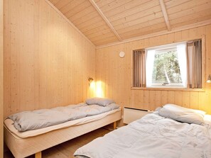 A room with wooden walls and a window with curtains. There are two beds with pillows and a bedside table.
