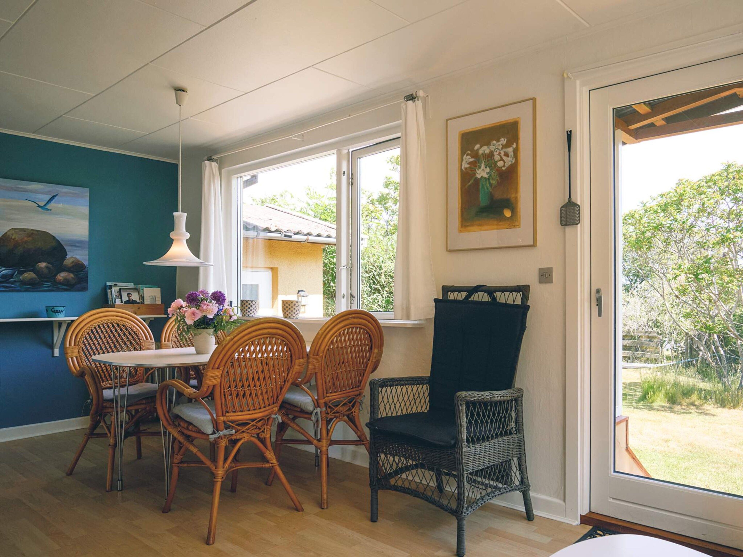 Un comedor con sillas de mimbre, una mesa y un florero con flores. Se ve una ventana con cortinas, un cuadro en la pared y vistas al exterior.