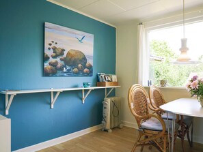 A room with a blue wall, a painting of a bird and rocks, a white shelf with books, a radiator, wicker chairs, and a window with a view of greenery.