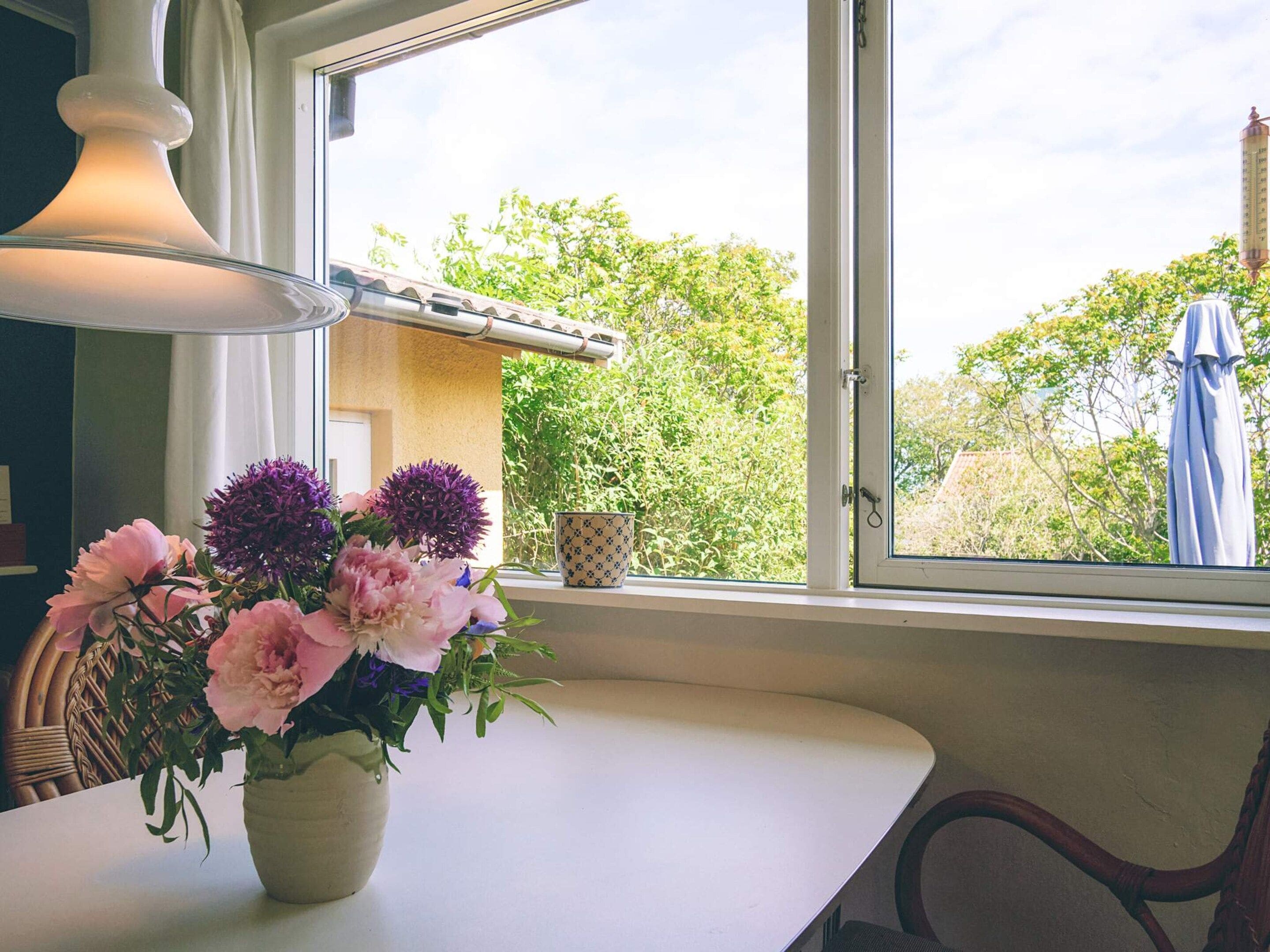 Una mesa con un florero, una ventana con vistas a follaje, y una lámpara.