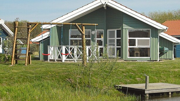 A house with a green exterior and white trim, a wooden deck, and a small dock extending into a body of water. - Holiday Home in Otterndorf (Otterndorf)