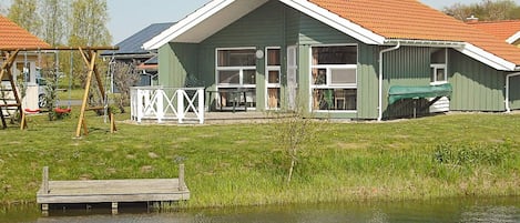 Una casa verde con techo inclinado, un muelle y un estanque con patos.