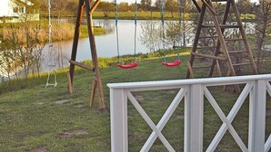 A wooden deck with a white railing, a swing set with red seats, and a calm body of water in the background. - Holiday Home in Otterndorf (Otterndorf)