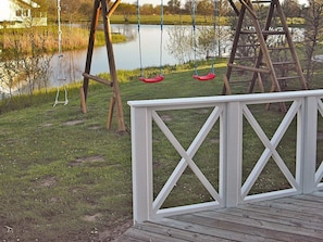 A wooden deck with a white railing, a swing set with red seats, and a calm body of water in the background. - Holiday Home in Otterndorf (Otterndorf)