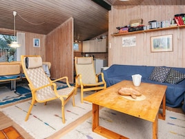 Una habitación acogedora con paredes de madera, un sofá azul, dos sillas de madera, una pequeña mesa con pan y una estantería con libros y DVD.