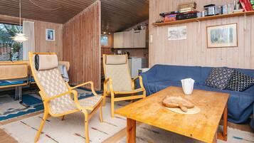 Une pièce chaleureuse avec des murs en bois, un canapé bleu, deux chaises en bois, une petite table avec du pain et une bibliothèque remplie de livres et de DVD.