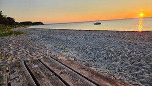 En træbro fører ned til en strand med en båd på vandet og en solnedgang i baggrunden.