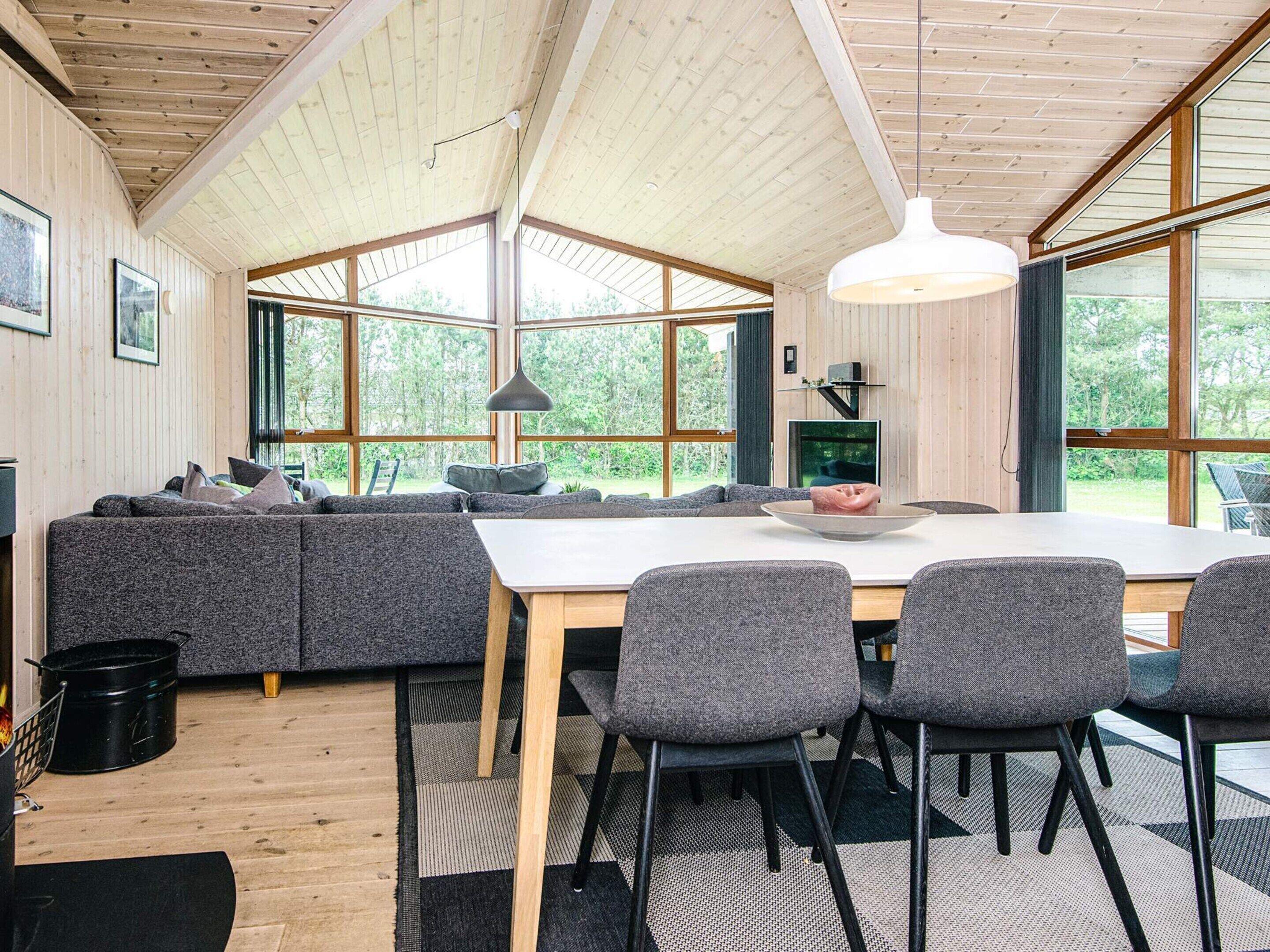 Un interior moderno con techo de madera, ventanas amplias, zona de comedor con mesa y sillas, y un rincón de estar acogedor con sofá y cojines.