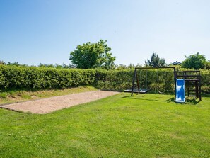 A playground with a slide and swings, surrounded by a hedge and a grassy area.