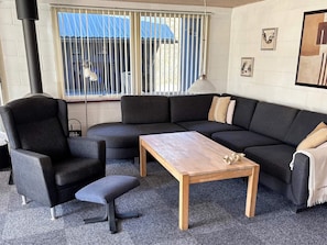 A living room with a sectional sofa, a wooden coffee table, a black armchair, and a window with blinds.