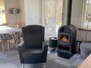 A modern living room with a black leather armchair, a built-in wood-burning stove, a dining table with chairs, and a window with vertical blinds. - 4 Star Holiday Home in Thyholm (Thyholm)