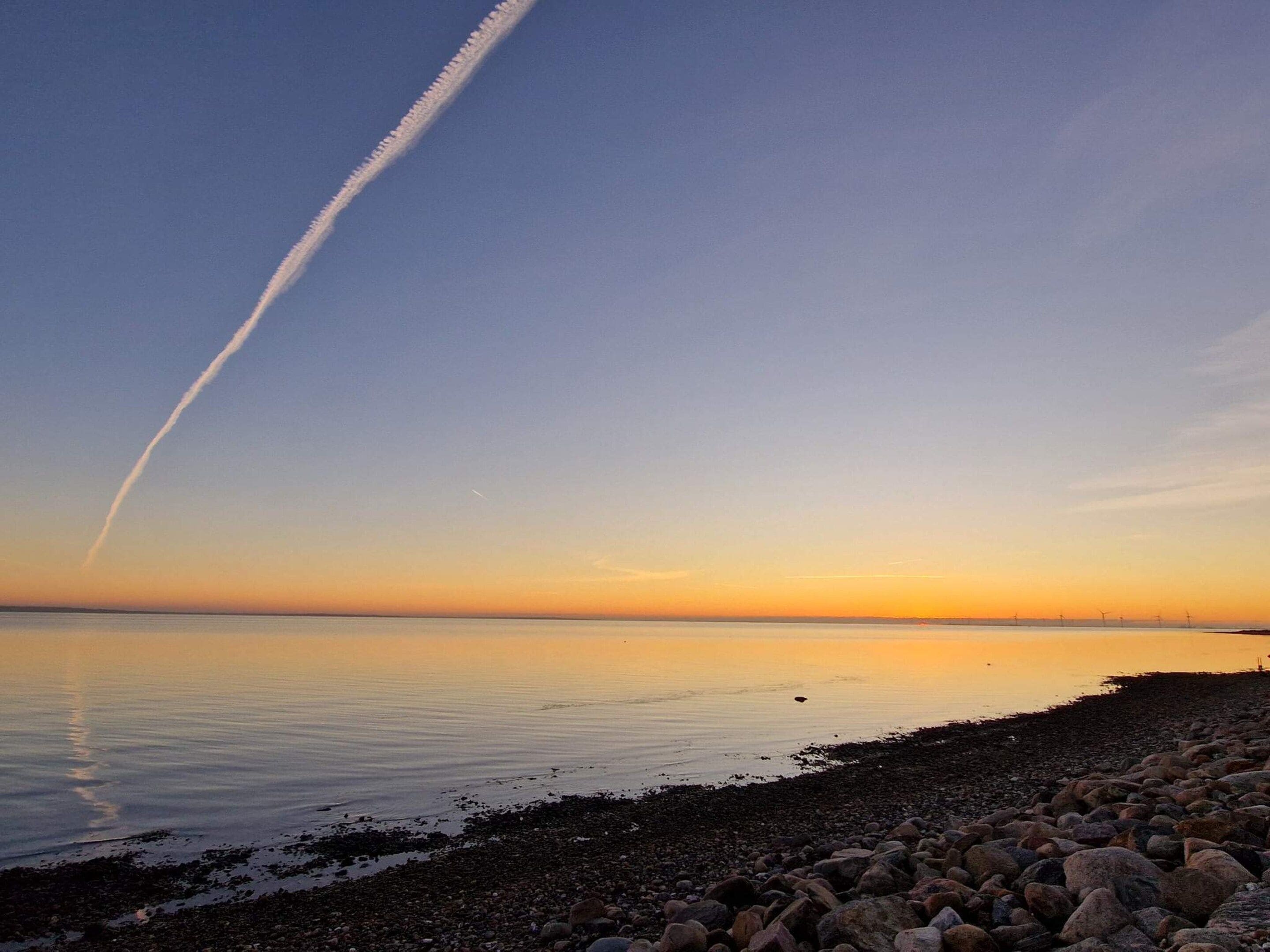 A calm beach with a clear sky, a visible contrail, and a rocky shoreline.