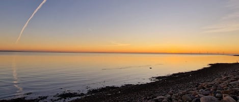 A calm beach with a clear sky, a visible contrail, and a rocky shoreline.