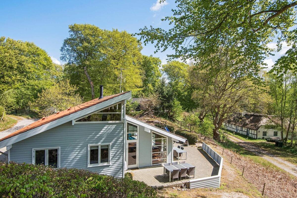 A two-story house with a sloped roof, a patio with outdoor furniture, and a surrounding wooded area.