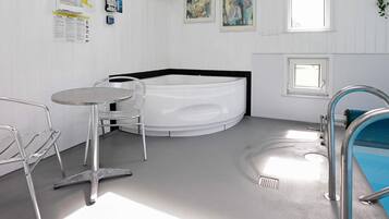 A modern bathroom with a bathtub, a small table, and a chair. There are two framed artworks on the wall and a window providing natural light.