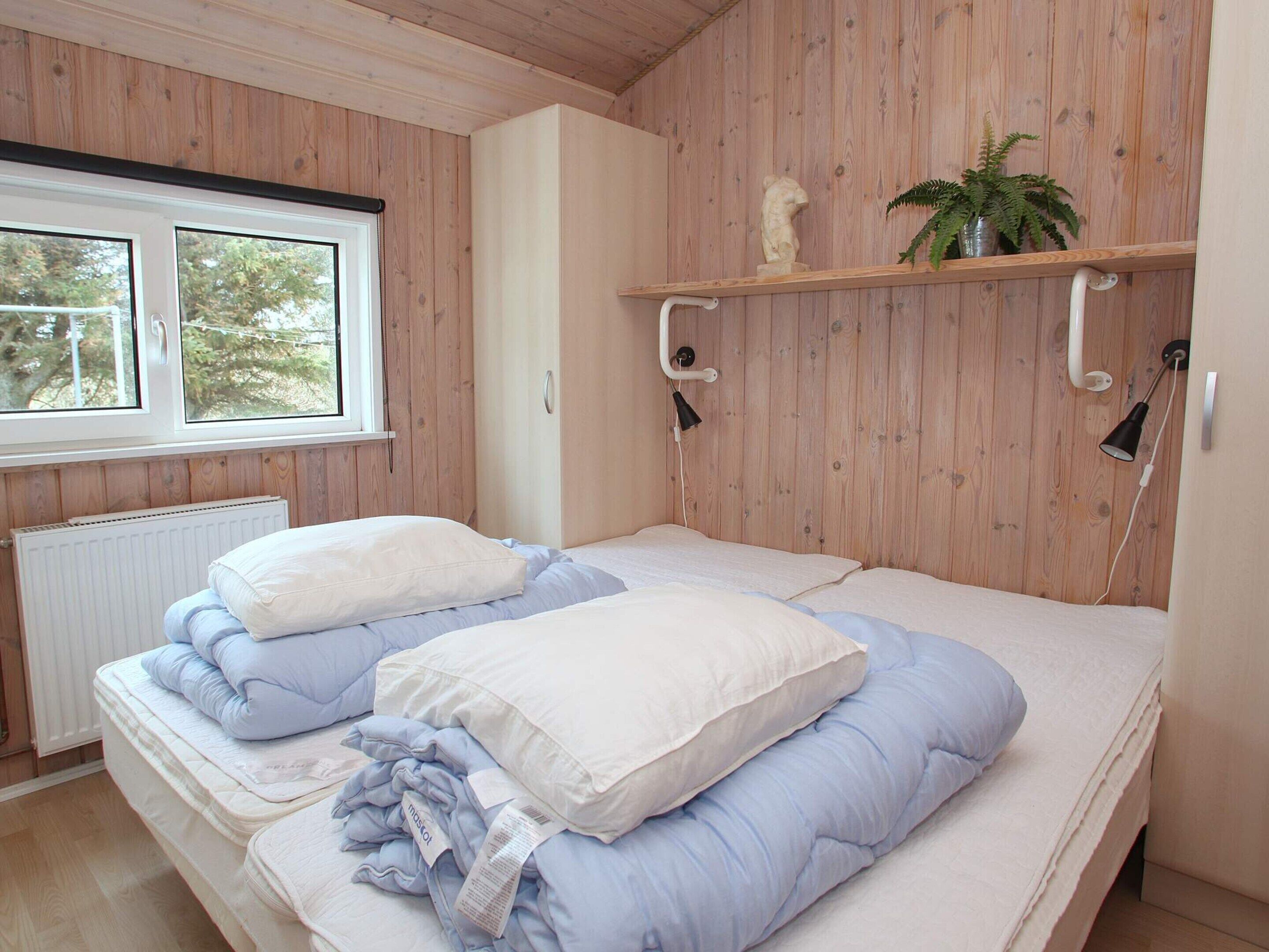 Een slaapkamer met een houten wand, een raam, een bed met kussens, een radiator en een plank met een plant.