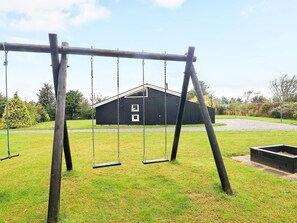 Eine Holzschaukel auf einer Rasenfläche, im Hintergrund ein schwarzes Gebäude.