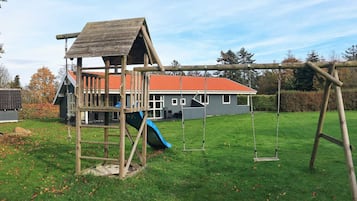 A wooden play structure with a slide and swings, a red-roofed building, and a grassy yard.