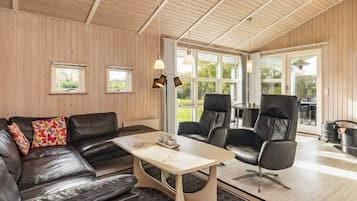Een moderne woonkamer met een leren bank, een salontafel en twee fauteuils. Grote ramen zorgen voor natuurlijk licht en uitzicht op het omliggende landschap.