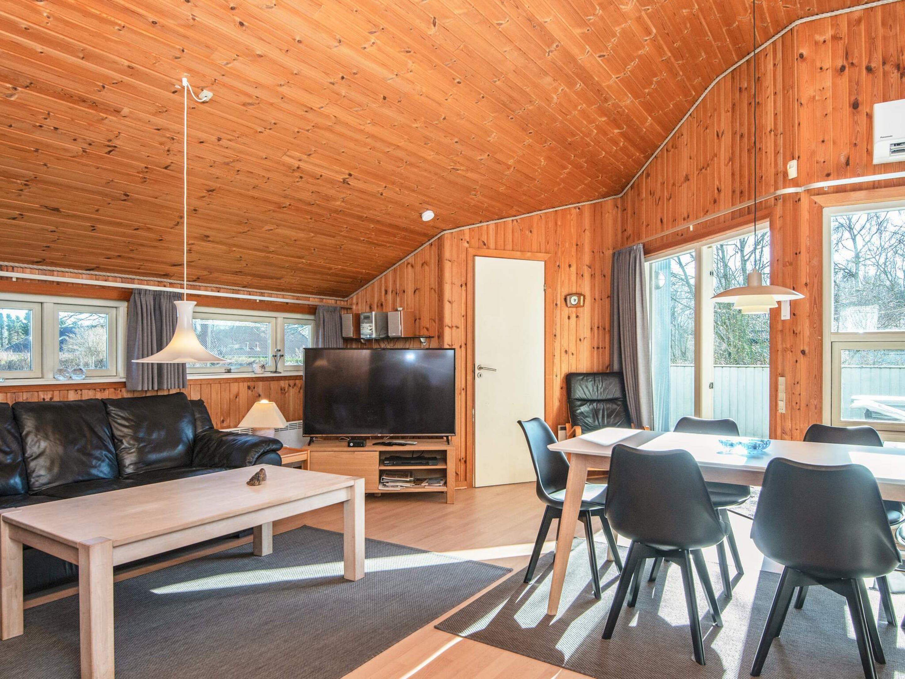 A cozy cabin interior with a wooden ceiling, a flat-screen TV, a leather sofa, a dining table with chairs, and a large window.