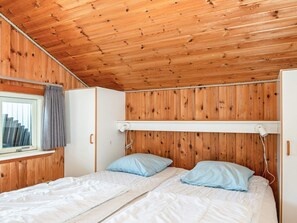 A room with wooden walls and a sloped ceiling, featuring a bed with two pillows and a window with curtains.