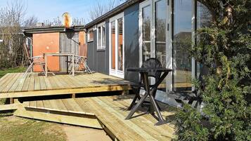 Eine Holzdielen-Terrasse mit Tisch und Stühlen, ein gläserner Anbau und ein kleines, oranges Gebäude.