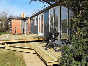 Eine Holzdielen-Terrasse mit Tisch und Stühlen, ein gläserner Anbau und ein kleines, oranges Gebäude.