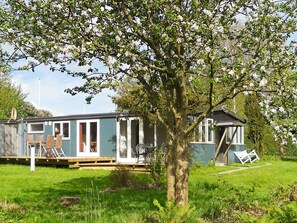 A mobile home with a deck and a small attached structure, surrounded by grass and trees.