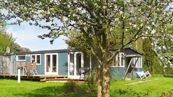 A mobile home with a deck and a small attached structure, surrounded by grass and trees.