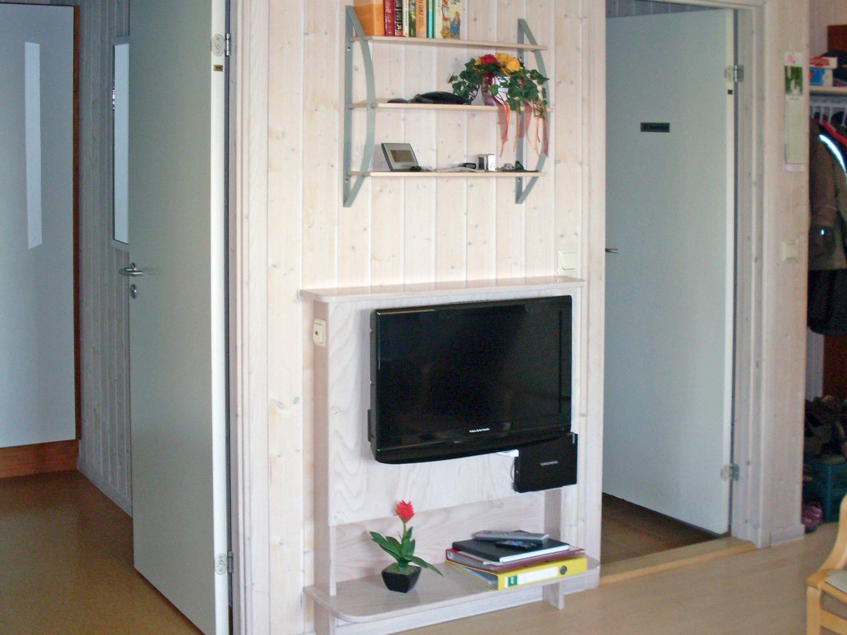Une pièce avec un téléviseur fixé au mur, un mur en bois, une petite étagère avec des livres et une fleur, et une porte sur le côté gauche.