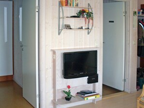 A room with a television mounted on a wooden wall, a small shelf with books and a flower, and a door to the left. - Holiday Home in Otterndorf (Otterndorf)