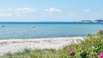 A beach with clear blue water, boats, and distant hills.