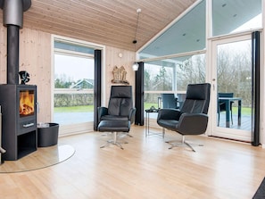 A modern living room with a wood-burning stove, two leather chairs, a glass coffee table, and large windows offering a view of the outdoors. - 5 Star Holiday Home in Hemmet (Hemmet)