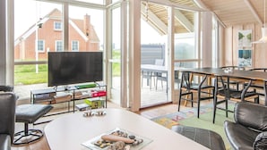 A modern living room with a large TV, comfortable seating, and a view of a brick house through large windows. - 7 Person Holiday Home in Harboore-by Traum (Harboore)