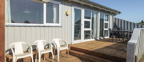 A wooden deck with white plastic chairs and a table, a sliding glass door, and a view of the surrounding area.