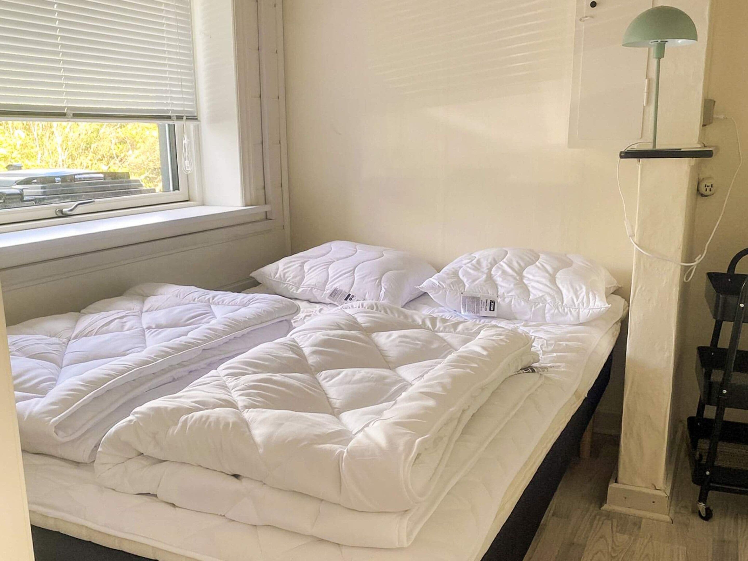 A single bed with a white duvet and pillows, a window with blinds, and a wall-mounted lamp.