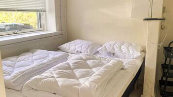 A single bed with a white duvet and pillows, a window with blinds, and a wall-mounted lamp.