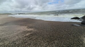 Une plage de galets, des vagues, un ciel nuageux et une éolienne à l’horizon.