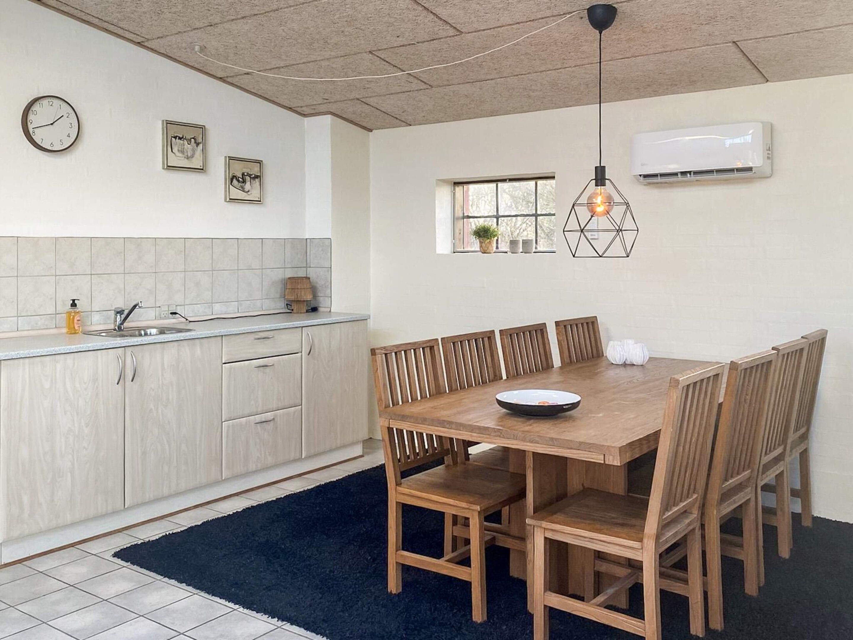 A kitchen with a dining area, featuring a wooden table and chairs, a clock, and a window with plants.