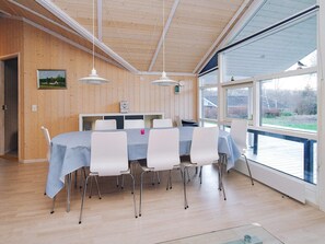 A dining area with a long table, white chairs, and pendant lights. There is a window with a view of the outdoors. - Cozy Holiday Home in AsnÃ¦s near Fishing (Asnaes)