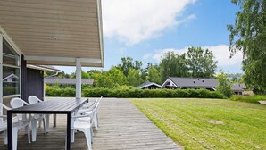 A wooden deck with a table and chairs, surrounded by a well-maintained lawn and a row of houses in the background. - Cozy Holiday Home in AsnÃ¦s near Fishing (Asnaes)